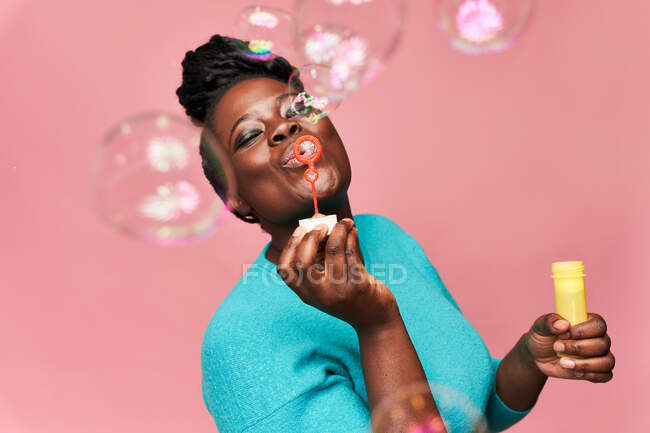 Heureuse femme afro-américaine avec les yeux fermés portant des vêtements bleus et soufflant des bulles de savon sur fond rose en studio — Photo de stock
