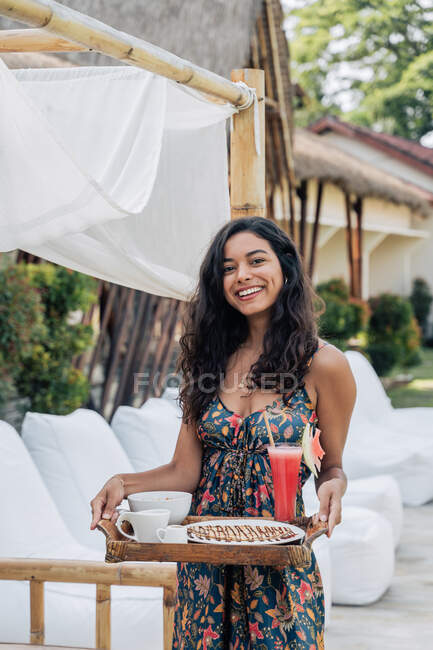Smiling female tourist in sundress holding tray with delicious breakfast while looking at camera during trip — Stock Photo