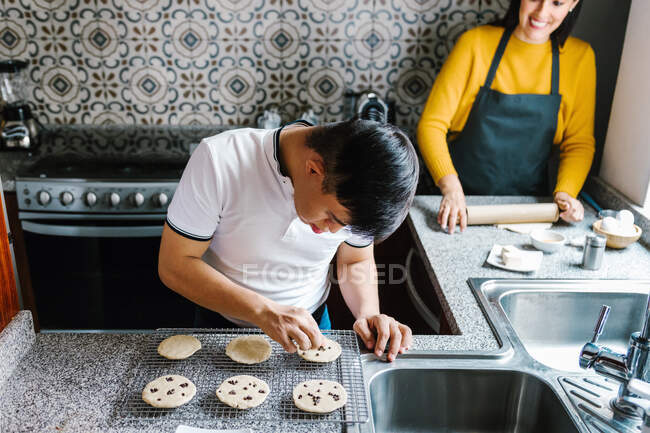Alto ângulo do adolescente latino com síndrome de Down decorando biscoitos crus com chips de chocolate enquanto cozinha na cozinha em casa — Fotografia de Stock
