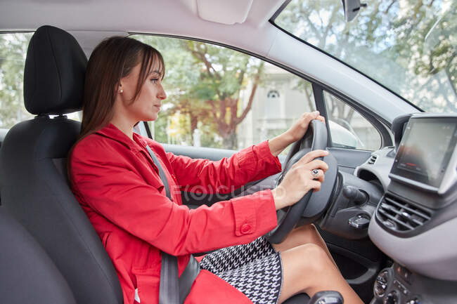 Through window side view of confident young female in red jacket driving modern automobile in city — Stock Photo