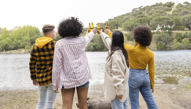Retrovisore anonimo di uomini e donne che si accarezzano bottiglie di birra e propongono brindisi mentre riposano sulla riva del fiume in natura — Foto stock