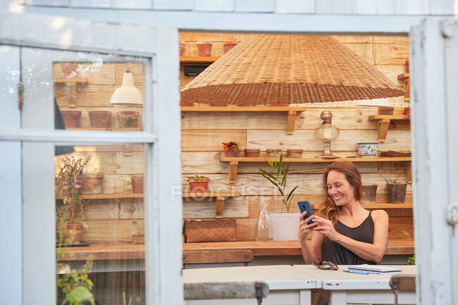 Konzentrierte Gärtnerin surft am Smartphone, während sie am Holztisch im Gewächshaus sitzt und arbeitet — Stockfoto