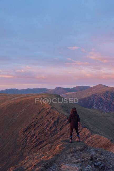 Vista posterior de caminante anónimo de pie en la colina rocosa en las tierras altas y disfrutar de la vista de la cresta de la montaña al atardecer en Gales - foto de stock