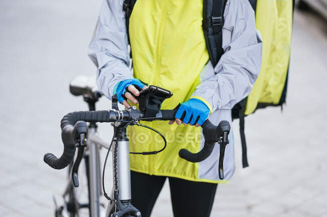 Crop view of anonymous young woman with thermal bag browsing smartphone with GPS map on bicycle handlebar while delivering food on city street — Stock Photo