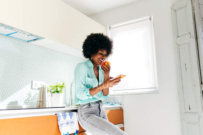 Знизу молода афроамериканка недбало носить яблуко і переглядає Інтернет на мобільному телефоні, стоячи вдома на кухні. — стокове фото