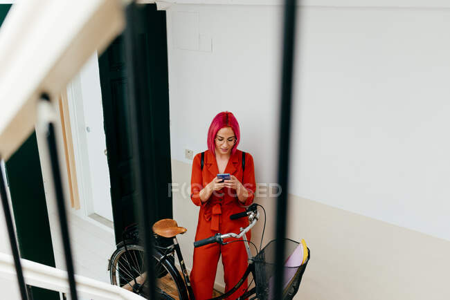 Зверху молода стильна жінка в червоному костюмі з рюкзаком за допомогою смартфона, стоячи з велосипедом на сходах — стокове фото