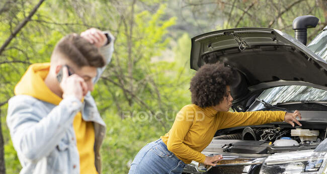African American female opening coolant container and repairing broken van near male making call to emergency service during road trip in countryside — Stock Photo