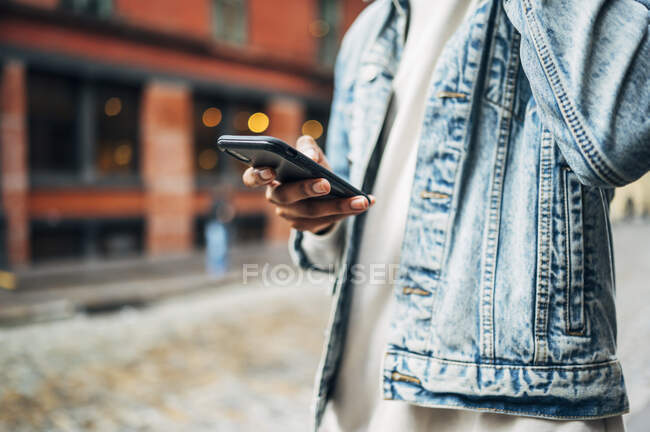 De baixo da colheita anônimo afro-americano cara em jeans jaqueta na moda surfar telefone celular moderno durante o passeio pela cidade — Fotografia de Stock