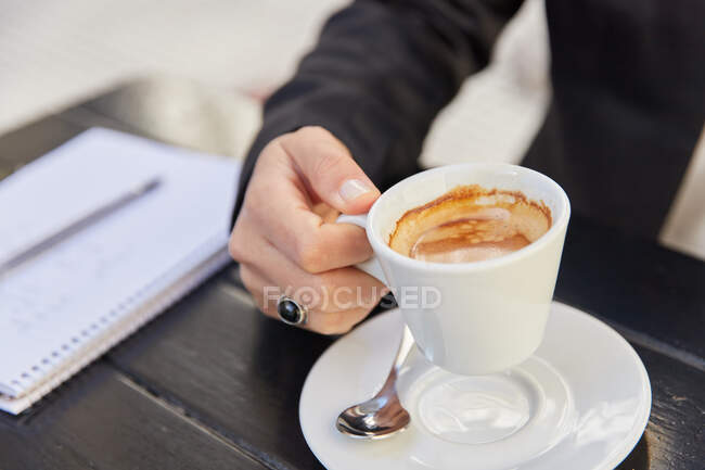 Cropped unrecognizable modern female entrepreneur drinking coffee while sitting at table with notebook urban outdoor cafeteria — Stock Photo