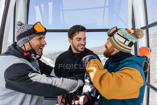 Alegres atletas masculinos en ropa deportiva hablando y mirándose el uno al otro en cabina de teleférico contra Sierra Nevada en España - foto de stock