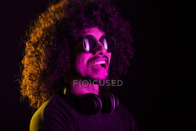 Positivo maschio latino dai capelli ricci in eleganti occhiali da sole e con cuffie sul collo distogliendo lo sguardo su sfondo nero in studio con luci al neon gialle e rosa — Foto stock