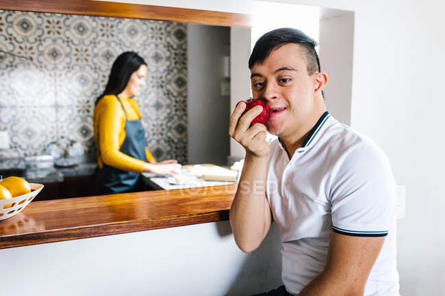 Content Latin teen boy with Down syndrome eating ripe red apple while sitting in kitchen with blurred mother and looking at camera — Stock Photo