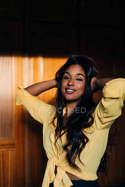 Молодая латиноамериканка в модной желтой рубашке смотрит в камеру, стоя с руками за головой в тени — стоковое фото