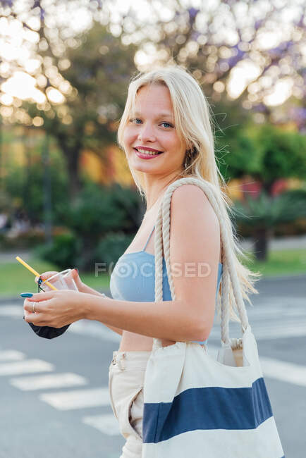 Seitenansicht einer fröhlichen Frau, die im Sommer mit kalter Limonade im Plastikbecher auf der Straße steht und in die Kamera blickt — Stockfoto