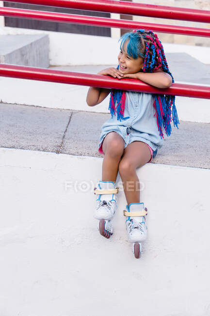 Fröhliches mexikanisches Kind mit hellen Zöpfen auf Rollschuhen sitzt, während es mit den Händen am Zaun lehnt und wegschaut — Stockfoto