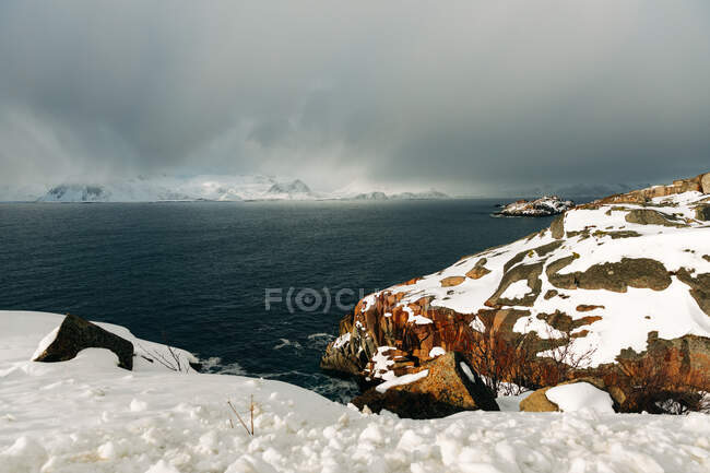 Schneebedeckte Felsbrocken und Bergrücken an der Küste in der Nähe von plätscherndem Meer gegen wolkenverhangenen Himmel im Winter auf den Lofoten, Norwegen — Stockfoto