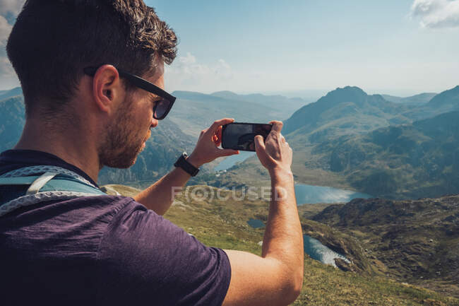 Vista laterale dell'esploratore maschio che scatta foto di montagne rocciose sul cellulare durante il trekking in estate in Galles — Foto stock