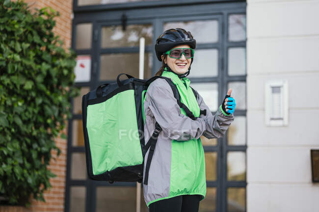 Mujer entrega feliz con bolsa térmica mirando a la cámara con sonrisa y haciendo gestos pulgar hacia arriba fuera del edificio de apartamentos - foto de stock