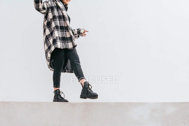 Donna irriconoscibile in cappotto a scacchi alla moda in equilibrio sul confine mentre cammina contro il muro grigio sulla strada — Foto stock