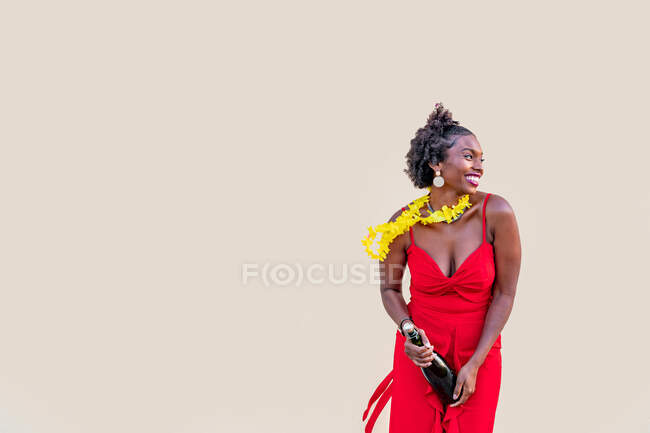 Delighted African American female in flower necklace wearing bright overall standing with bottle of champagne against white background — Stock Photo