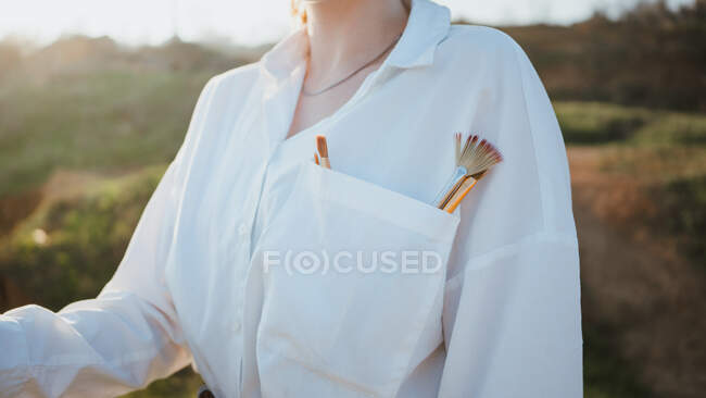 Cultiver jeune femme debout sur la côte herbeuse près du sable et de l'océan par temps ensoleillé avec des pinceaux sur la poche — Photo de stock