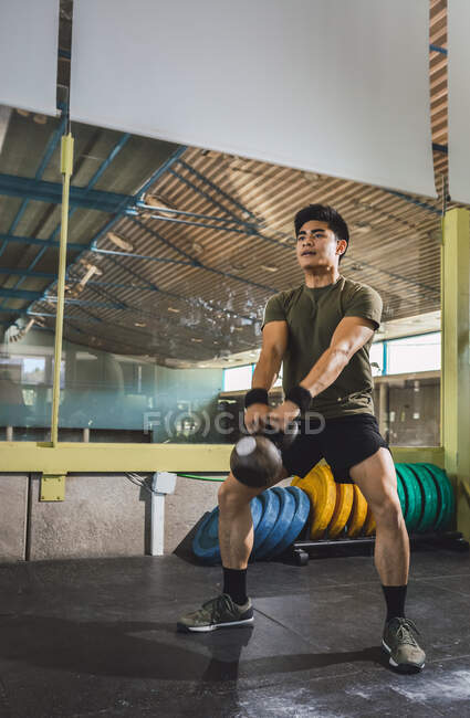 Asian man training shoulders and arms with heavy kettlebells in gym during functional workout and looking away — Stock Photo