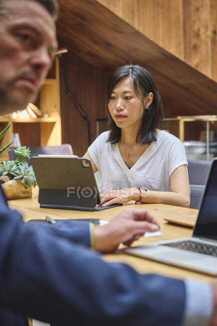 Азиатка, работающая в своем ноутбуке в офисе с другим коллегой — стоковое фото