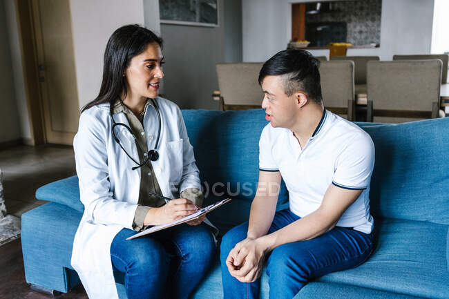 Mujer alegre practicante en bata médica hablando con un adolescente con síndrome de Down y tomando notas en el portapapeles durante la cita en casa - foto de stock