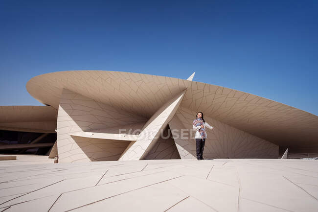 Mulher asiática feliz sorrindo enquanto está de pé contra o magnífico exterior do edifício do Museu Nacional com arquitetura incomum consistindo de muitas superfícies listradas circulares no Qatar — Fotografia de Stock