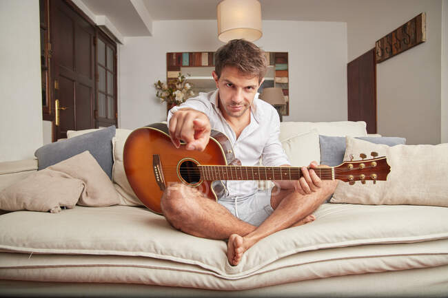 Взрослый мужчина в повседневной одежде сидит на диване с акустической гитарой в руках, указывая пальцем на камеру в светлой гостиной и глядя в камеру — стоковое фото