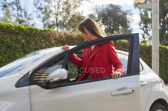 Модная молодая женщина-водитель в стильной красной куртке садится в современный серый автомобиль, припаркованный возле зеленых растений весной — стоковое фото