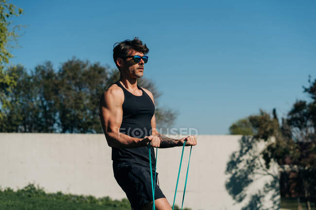 Strong male athlete in sportswear and sunglasses exercising with elastic band on lawn in sunlight — Stock Photo