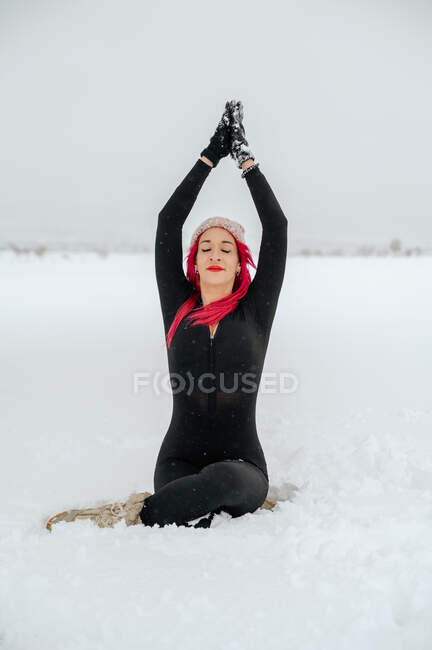 Conteúdo feminino com cabelo rosa praticando ioga em Gomukhasana e alongamento pernas enquanto sentado com gesto Namaste no campo de inverno nevado — Fotografia de Stock