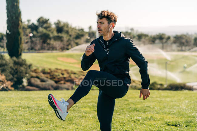 Дорослий спортсмен-чоловік у спортивному одязі, що піднімає ногу і з нетерпінням чекає під час тренувань на газоні на сонячному світлі — стокове фото
