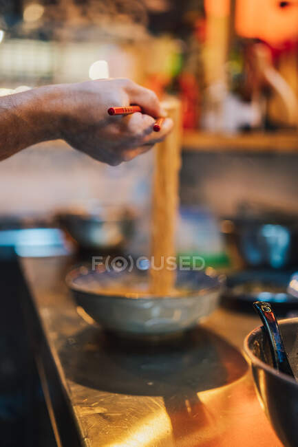 Crop hand of faceless chef with wooden chopsticks holding noodle above bowl on blurred background in ramen bar — Stock Photo
