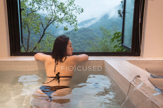 Jeune femme ethnique détendue en maillot de bain couchée dans un bain d'onsen japonais dans une station thermale à côté d'une fenêtre avec vue sur les montagnes et les arbres verts — Photo de stock