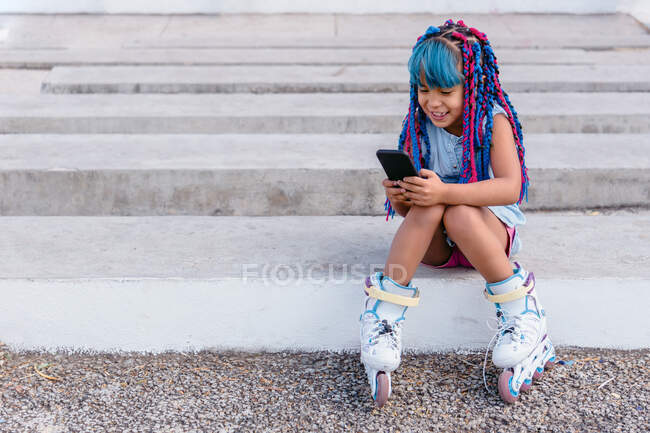 Criança mexicana alegre em patins com tranças coloridas navegando na internet no celular enquanto descansa nas escadas ao ar livre — Fotografia de Stock