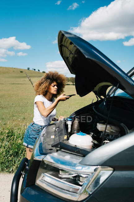 Joven mujer étnica con cabello afro llamando para reparar el servicio en el teléfono inteligente mientras está de pie cerca de autocaravana con capucha abierta en la carretera rural en el día de verano - foto de stock