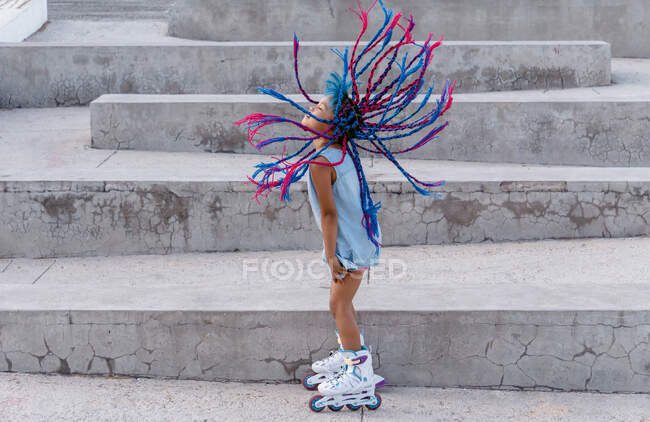 Side view of ethnic child in roller skates with flying colorful braids standing on staircase — Stock Photo