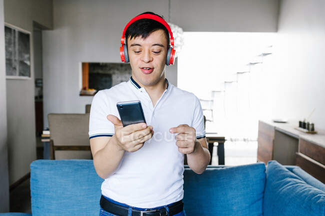 Приємний латиноамериканський підліток у навушниках на телефоні, стоячи біля дивана вдома і дивлячись на камеру. — стокове фото