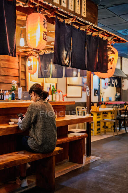 Asian lady in casual sweater using mobile phone at counter in traditional ramen bar — Stock Photo