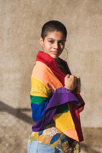 Молода бісексуальна етнічна жінка з різнокольоровим прапором дивиться на камеру і представляє символи ЛГБТК в сонячний день — стокове фото