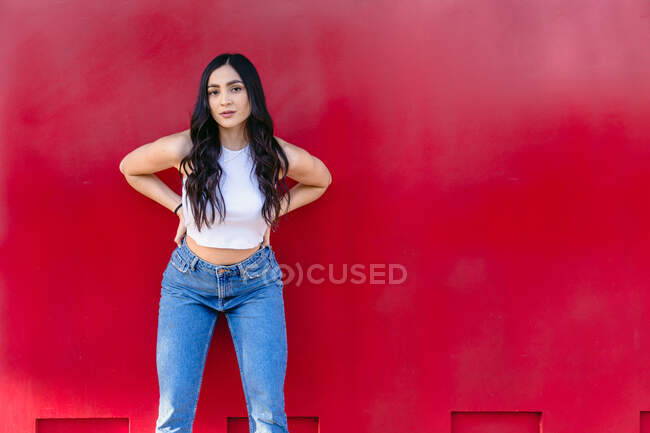 Unemotional female teenager in casual outfit looking at camera standing against red wall — Stock Photo