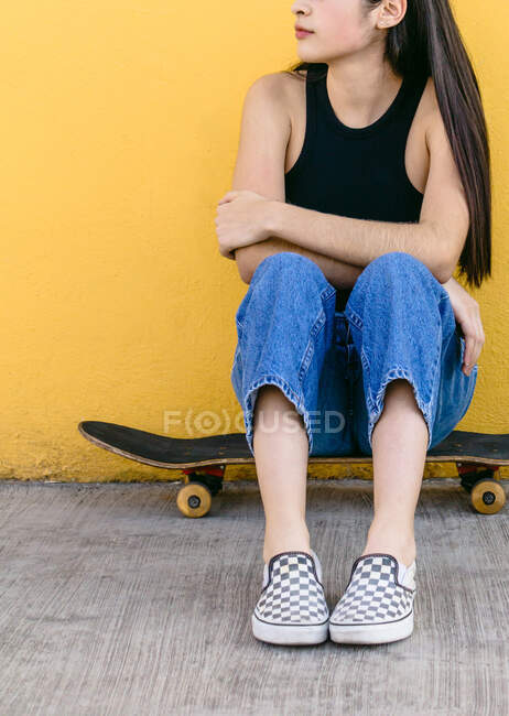 Cropped unrecognizable young dreamy female skateboarder in casual outfit sitting on board on walkway — Stock Photo