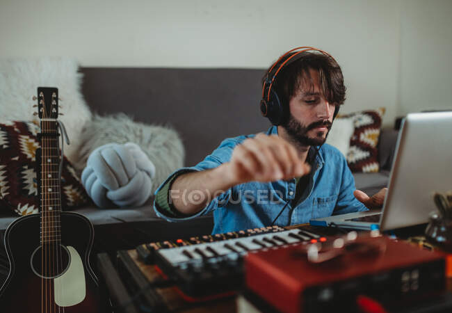 Young man in headphones using synthesizer and laptop at table at home — Stock Photo