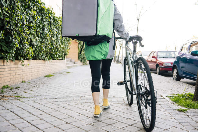 Crop mensajero femenino con bolsa térmica en blanco caminando cerca de la bicicleta en el pavimento en la calle de la ciudad - foto de stock