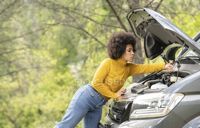 Seitenansicht einer Afroamerikanerin, die Kühlmittelbehälter öffnet und kaputte Lieferwagen während einer Autofahrt in der Landschaft repariert — Stockfoto
