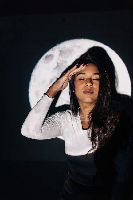 Jovem fêmea hispânica com olhos fechados tocando a cabeça enquanto descansa sob projeção lunar à noite — Fotografia de Stock