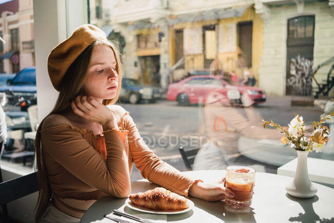 Французская женщина в берете сидит за столом в кафе с ароматным стаканом кофе и свежеиспечённым круассаном — стоковое фото