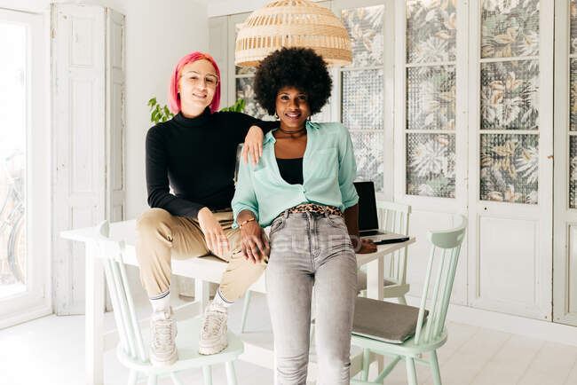 Alegres jovens amigos femininos multirraciais elegantes olhando para a câmera enquanto sentados juntos na mesa na sala de luz — Fotografia de Stock